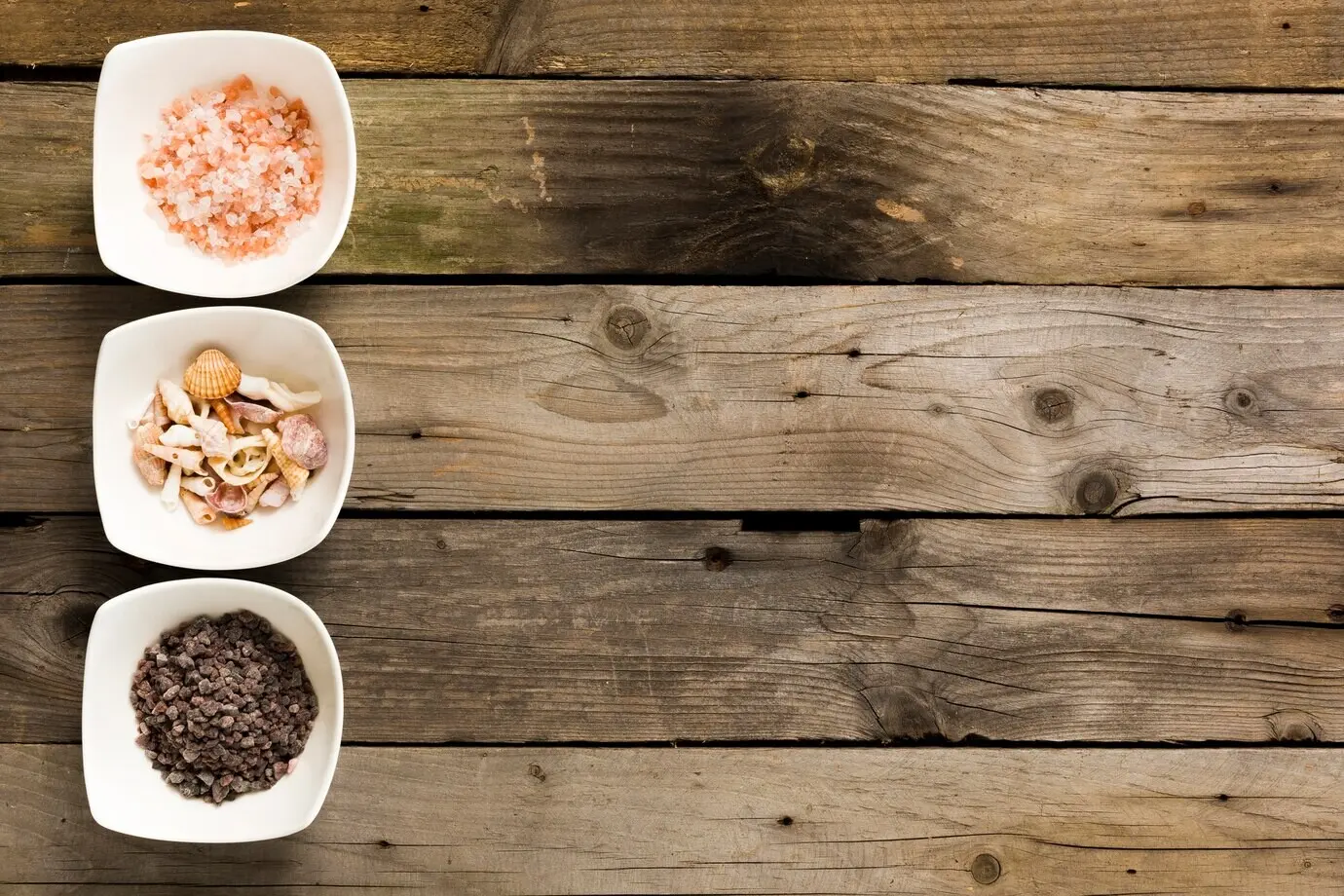A bowl of pink and black salt with seashells on a wooden table.